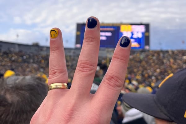 A hand holding up a three on the field after Michigan beat OSU. Two nails are painted blue, a 3rd yellow with the JJ smile.